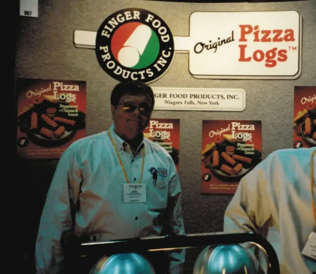 Robert Cordova, founder of Finger Food Products, LLC, in front of display of Original Pizza Logs Logo, Finger Food Products logo, and information about Original Pizza Logs® Products.